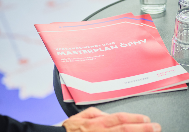 Eine Hand liegt auf einem Tisch. Daneben liegt ein rotes Magazin. Darauf stehen die Wörter „Verkehrswende 2030“, „Masterplan ÖPNV“ und „Vestische“. Auf dem Tisch steht auch ein Glas Wasser.
